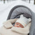 A sleeping newborn baby wearing a white knit hat with ear-like details, nestled inside a gray stroller with a cream fleece liner, set against a snowy outdoor background.