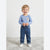A smiling toddler stands on a light gray rug wearing a light blue long-sleeve top with a small multicolor animal graphic on the chest, blue jeans, and white shoes.