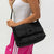 A woman wearing a white tank top and pink athletic pants carries a black quilted puffer shoulder bag against a plain light gray background.