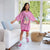 A young girl stands in a bright living room wearing an oversized pink graphic T-shirt dress with 'ARMY CORE SUMMER' text and a gift box illustration. She is wearing pink sneakers and white socks, with a white sofa, blue pillows, and a wicker basket chair nearby.