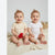 Two smiling babies sitting side by side on a cream blanket. The baby on the left wears a pale yellow short-sleeve bodysuit with tiny red polka dots and holds a red heart-shaped toy.