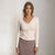 A woman modeling a fitted long-sleeve V-neck knit top in a light cream color with a button placket, paired with a sparkly mauve skirt, against a plain light gray studio background.