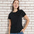 A woman wearing a plain black short-sleeve crewneck t-shirt, standing against a white brick wall.