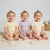 Three smiling babies sitting side by side on a cream knit blanket, wearing short-sleeve bodysuits in pale yellow, lavender, and cream. A woven basket with plush toys is visible in the background against a light gray studio backdrop.