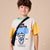 A young boy with brown hair smiling, wearing a color-blocked t-shirt with yellow sleeves, a cream body, and a black graphic print. He has a black crossbody bag slung over his shoulder, set against a solid beige background.