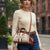 A woman wearing a cream ribbed sweater and blue jeans carries a beige and brown dome-shaped crossbody bag with white stripes and the brand name 'bebe' printed on the front, set against a blurred city street background.