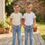 Two young girls standing side-by-side on a wooden deck outdoors, wearing matching light-wash blue jeans and short-sleeve t-shirts. The girl on the left wears a cream t-shirt with small brown polka dots, and the girl on the right wears a white t-shirt with small red dots.