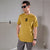 A man wearing a mustard yellow t-shirt with a black graphic logo on the chest, styled with gray cargo pants, black sunglasses, and a black watch, standing against a minimalist gray wall.