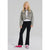 A young girl with blonde hair stands against a light gray studio background wearing a cropped, faded gray denim jacket over a white top, black flared pants, and pink sneakers.