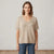 A woman wearing a short-sleeve V-neck knit sweater in a light beige color, paired with blue jeans, standing against a neutral off-white wall.