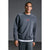A man wearing a dark gray crewneck sweatshirt with a small red rectangular logo patch on the chest, layered over a white undershirt and gray sweatpants, set against a dark gray studio background.