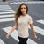 A woman walking on a city crosswalk wearing a light beige crewneck t-shirt with a small chest pocket and black pants. The t-shirt features a cursive graphic that reads 'Find your Balance' with small hearts.