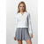A blonde model wearing a white long-sleeve polo sweater and a gray pleated mini skirt, standing against a light gray studio background.