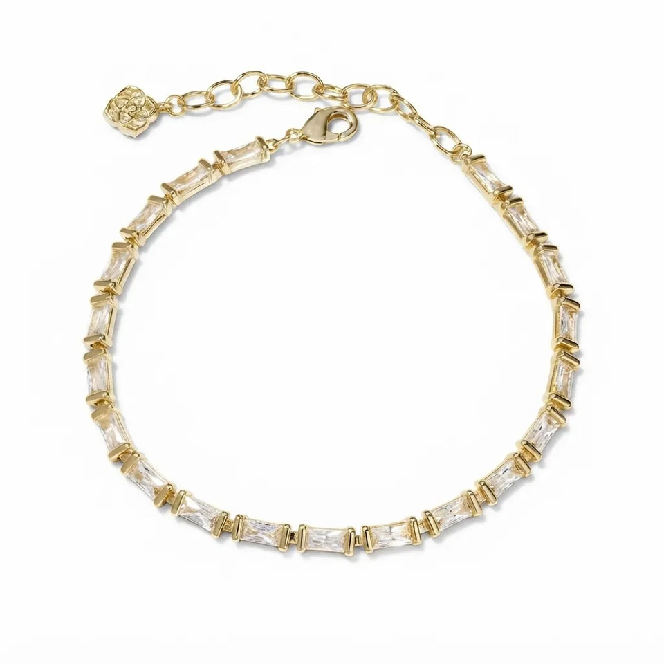 A gold chain bracelet featuring a row of clear baguette-cut crystals, a lobster clasp, and an adjustable extender chain with a decorative flower-shaped tag, shown on a plain white background.