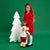 A young girl in a red dress and white tights stands next to a white Christmas tree decoration and a plush mouse toy wearing a white sweater, red pants, and a red-and-white plaid scarf, against a solid green studio background.