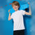A smiling young boy with light brown hair stands against a bright blue background, holding a light blue penny board over his shoulder. He is wearing a plain white short-sleeve t-shirt and black shorts.