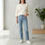 A woman wearing light-wash blue straight-leg jeans, a cream-colored knit sweater, and white sneakers, standing in a bright, modern room with a concrete pillar and a potted plant.