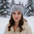 A woman wearing a light gray twisted knit headband adorned with small white pearl-like beads, photographed outdoors in a snowy setting with blurred evergreen trees in the background.