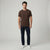 A man modeling a dark brown short-sleeve crewneck t-shirt, paired with dark navy jeans and white sneakers, standing against a light gray studio background.