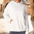 A person wearing an oversized white pullover hoodie with a kangaroo pocket, standing in a sunlit outdoor alley with blurred stone buildings in the background.