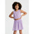 A young girl with curly dark hair wearing a light purple two-piece set consisting of a sleeveless cropped top and a matching flared skirt, posed against a light gray studio background.