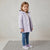 A young girl stands on a textured gray rug wearing a light lavender quilted hooded jacket, blue jeans, and matching lavender sneakers against a neutral studio backdrop.