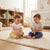 Two toddlers sitting on a chunky cream knit rug in a bright nursery, playing with wooden blocks and a plush elephant toy. One child wears a white shirt and yellow pants, and the other wears a light gray shirt and navy pants.
