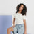 A young woman with curly dark hair wearing a fitted off-white short-sleeve t-shirt and light-wash denim shorts, posing against a white wall with a blue geometric block.