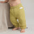 A baby wearing textured olive green gauze pants, standing barefoot next to a beige knit blanket.