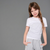 A young girl with long light brown hair stands against a solid gray studio background, wearing a white short-sleeve crewneck t-shirt and light gray sweatpants.