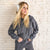 A woman with blonde hair wearing a dark gray zip-up hoodie and matching sweatpants, standing against a white brick wall and wearing fluffy white earmuffs.