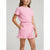 A young girl wearing a light pink two-piece outfit consisting of a short-sleeve crewneck t-shirt and a matching ruffled mini skirt, shown against a plain light gray studio background.