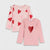 Two pink long-sleeve children's tops with ruffle details, displayed on a light gray background. The top on the left has scattered red and pink hearts, while the top on the right features a large red glitter heart on the chest.