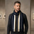 A man wearing a dark charcoal gray sweater and a thick knit scarf in navy blue, cream, and white stripes, standing in a modern indoor setting with a textured gray wall background.