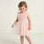 A toddler girl wearing a sleeveless light pink dress with a textured knit bodice, ruffled shoulder straps, and a layered tulle skirt with white floral embroidery, standing against a plain off-white studio background.