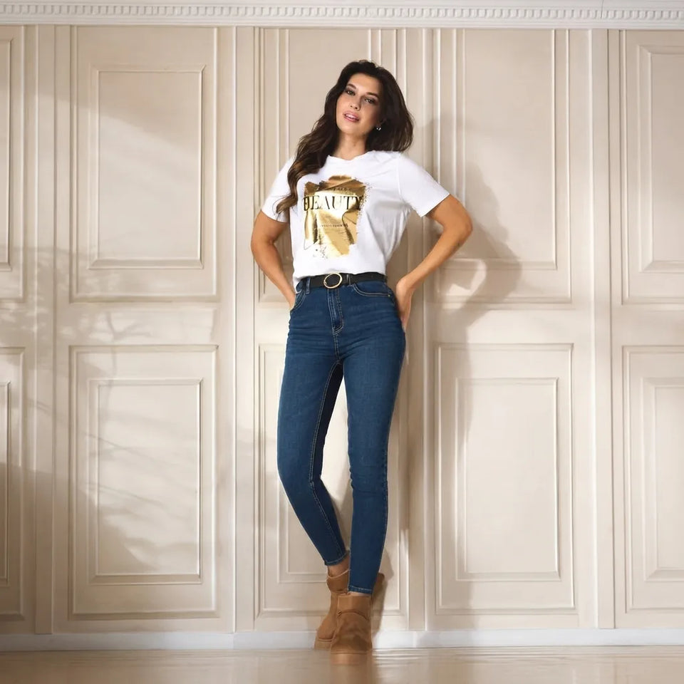 A woman stands against a cream paneled wall wearing a white graphic T-shirt with a gold print, dark blue skinny jeans, a black belt, and tan ankle boots.