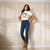 A woman stands against a cream paneled wall wearing a white graphic T-shirt with a gold print, dark blue skinny jeans, a black belt, and tan ankle boots.
