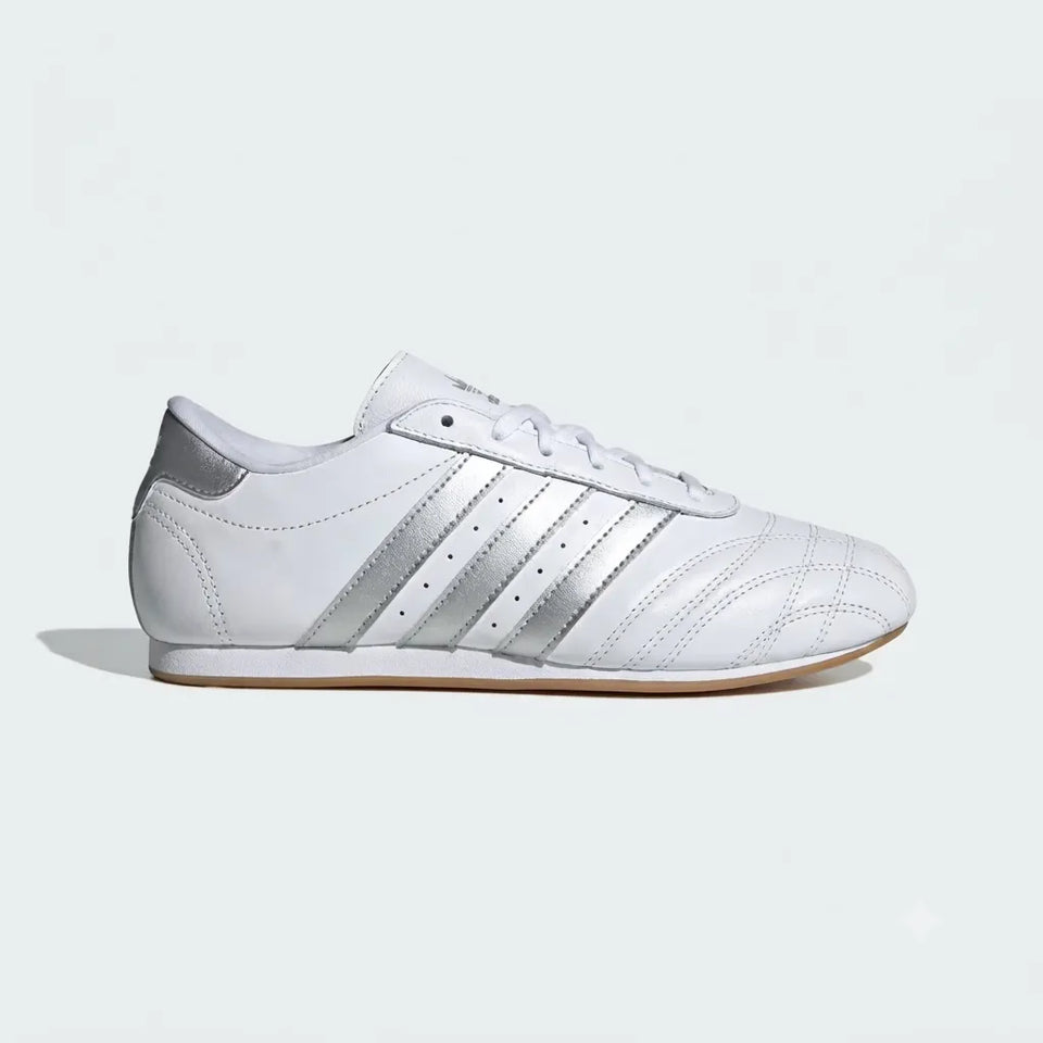 A white leather low-top sneaker with metallic silver stripes on the side, white laces, and a gum-colored outsole, shown in profile against a light gray background.