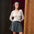 A woman models a cream ribbed long-sleeve wrap top paired with a gray pleated mini skirt and a matching gray headband with a white letter 'P'.