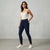 A woman modeling a pair of dark navy blue skinny jeans, paired with a cream camisole top and white sneakers, standing against a textured light gray studio wall.