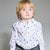 A toddler with blonde hair and blue eyes wearing a white long-sleeve button-up shirt with a festive pattern of green pine trees, brown reindeer, and candy canes, paired with gray pants, photographed against a neutral gray background.