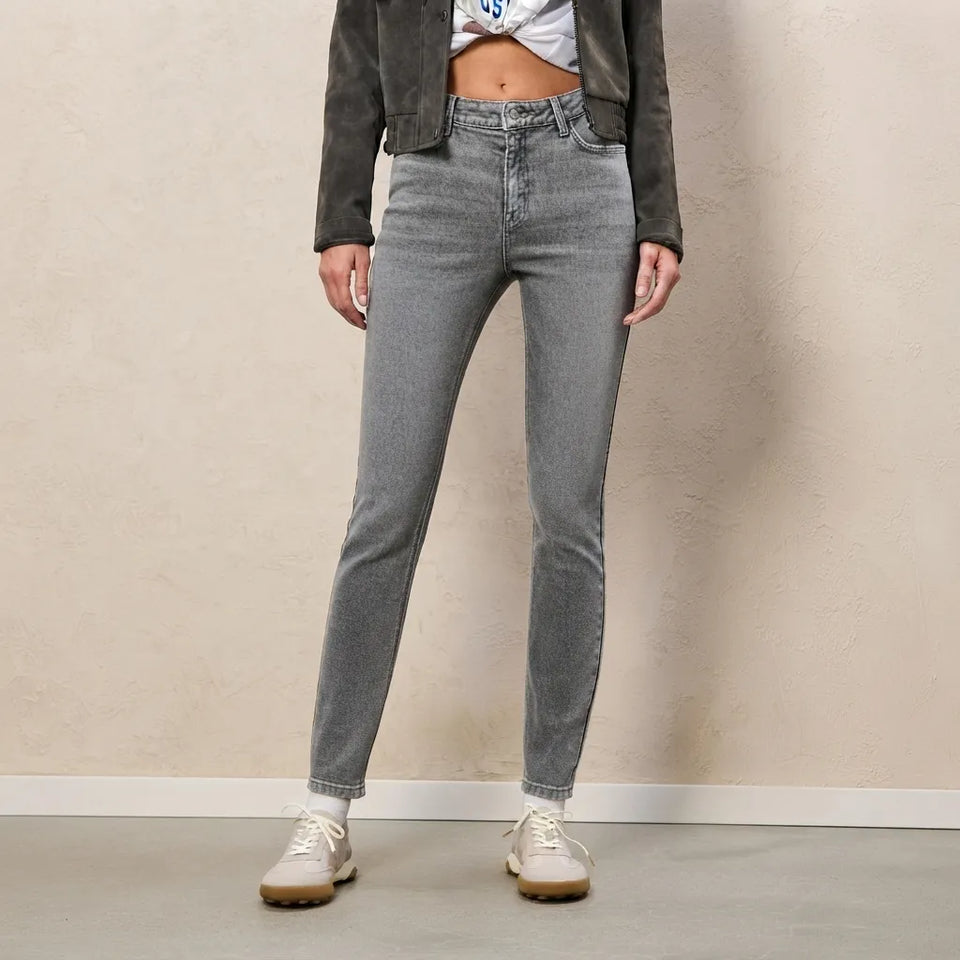 A model wearing gray slim-fit jeans, a dark gray suede jacket, a white crop top, and off-white sneakers, standing against a textured beige wall.