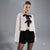 A model wearing a white textured boucle cropped jacket with a black bow at the neckline, paired with black sequined shorts, against a solid gray studio background.