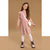 A young girl with long brown hair stands against a solid tan studio background wearing a dusty pink long-sleeve dress with ruffle details, white tights, pink lace-up boots, and a small white bunny-shaped crossbody bag.