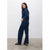 A woman modeling a dark blue denim set consisting of a long-sleeve collared shirt and matching wide-leg jeans, standing in a studio with a light gray wall and concrete floor.