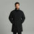 A man wearing a black mid-length puffer coat with a high stand-up collar, standing against a solid gray studio background.