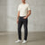 A man wearing dark charcoal slim-fit jeans, a cream t-shirt, and white sneakers, standing in a studio with a textured concrete wall and floor.