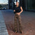 A woman stands on a red brick sidewalk in front of a modern glass building, wearing leopard-print wide-leg pants, a black sleeveless crop top, and black sandals.