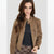A woman wearing a brown suede moto jacket with a buttoned collar, zippered front, and zippered chest pocket, styled over a black top and a brown plaid skirt, set against a plain light gray background.