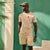 A man wearing a beige short-sleeve t-shirt and matching shorts with a green cap, standing against a teal wall with architectural columns.